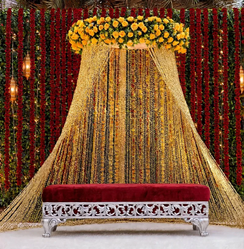Gold Curtain Mehndi Stage: Gold string curtain canopy with a yellow flower crown and a red velvet bench, in front of red and green flower strings, GuleGul Flowers Lahore
