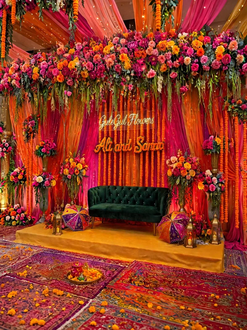 Orange and Pink Mehndi Stage: Orange and pink drapes with genda strings, a flower top, a green sofa and colourful rugs, GuleGul Flowers Lahore